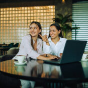 Two cheerful girls are sitting in a cafeteria with a laptop, pointing at people and gossiping. A girl is typing on a laptop and looking at something in a cafe while her friend is pointing.