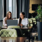 Two cheerful women are sitting in a cafe and smiling at the laptop while using credit card for online purchase. Happy friends are sitting in the cafeteria and purchasing online by using credit card.